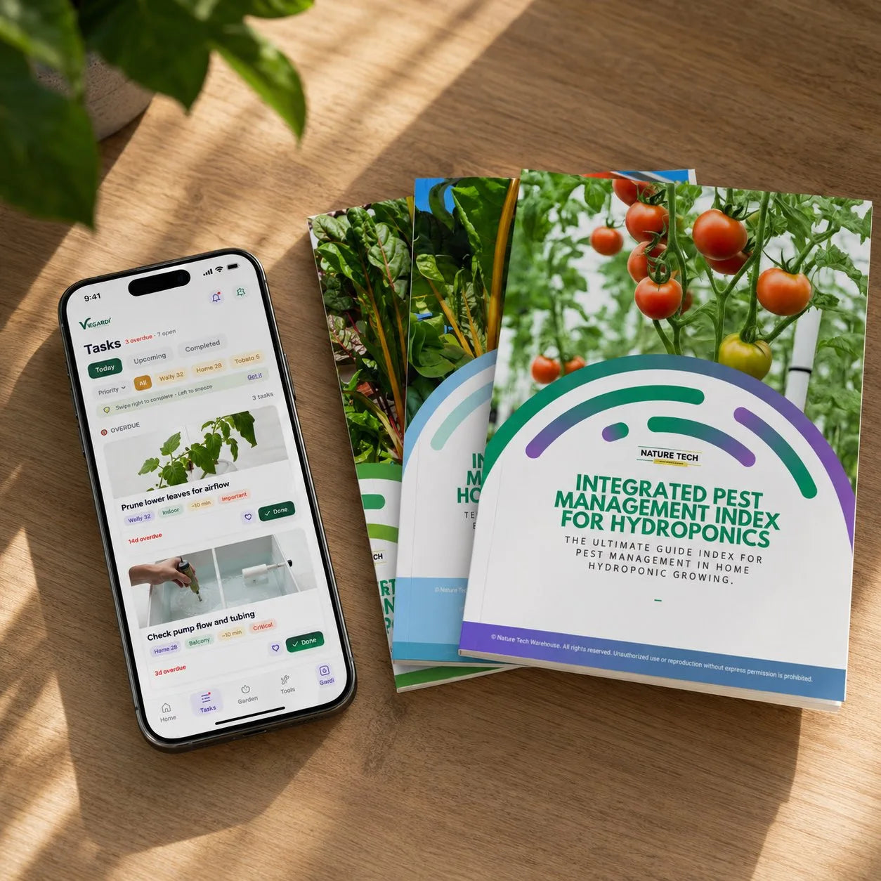 A phone showing the Vegardi Tasks screen alongside a stack of NatureTech printed guides on a wooden surface, with morning light and a hint of green foliage in the background